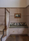 A built-in bench painted a dusty pink has two drawers of storage beneath and an olive green cushion on top, along with a cylindrical pillow in a painting-like forest print. To the left, the staircase can be seen.