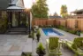An image of a lap pool next to this remodeled, single-family home. The pool is bright blue, with a flagstone patio and surrounding plants. There are two green lounge chairs of wood, and a step to the left up to a flagstone patio near the home.