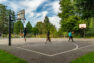 Four kids play basketball on a paved basketball court surrounded by grass on two sides, a wood on another, and a pickleball court on the fourth. There are two backboards and hoops. One child is attempting to make a basket.