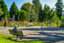 An outdoor, paved pickleball court with a single net. In the foreground, a park visitor observes a game between two people from a bench. Beyond is enclosed garden space and the park’s play area.