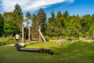 A play area for children at a public park. The play area has turf ground cover, a heavy lumber play structure to the right with a walking bridge and a climbing net, and a swing set to the right.