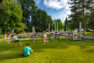 Kids run and play in front of an on a large, heavy timber play structure. There is a basketball court beyond the play area, and the whole park is surrounded by large, established trees.