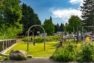 A public park play area with a short, zip line structure to the left with a child riding the line. Another child waits their turn, while other kids play on the heavy timber play structure to the left.