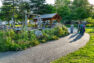 A group of four women walks along a paved path through the park. To the right of the path is a garden full of flowering plants. Beyond the garden is a covered picnic area and a play structure of heavy timber.