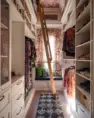 The primary closet in a remodeled, single-family home featuring white built-in storage on both sides and a ladder for accessing the higher shelves. At the back is a window, treated with a shade in the same floral pattern as the room’s multicolored wallpaper.