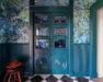 A teal-blue foyer and mudroom with antique doors from France. The doors are painted teal-blue like the room’s trim and wainscot. The floor is checkered white and black marble and the walls are covered in an abstract blue, green, and purple wallpaper.