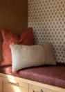 A close up of a built-in cabinet’s reading nook. The seat is upholstered in a dusty plum fabric that looks slightly textured. There is a terracotta colored square pillow on the bench, along with a rectangle shaped tan pillow. The back of the nook is covered in an wallpaper with a geometric flower pattern in neutral brown and white.