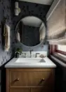 A straight on photograph of a powder room vanity, sink, and mirror. The room is narrow and the vanity take up the width of the wall, with a round mirror above. There is a small sconce to the left of the mirror and a window to the right.