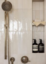 A close up image of a shower, including a polished nickel shower head and adjacent shower niche for storing products. The shower niche has two shelf levels. The shower’s tile wall is tiled in ceramic tiles oriented vertically. At the level of the shower head, the tile transitions from a white gloss to a white matte.