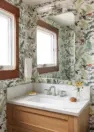 A bathroom decorated with white wallpaper with a fern and mushroom pattern in red and green. The bathroom vanity has one since and a white oak cabinet. Above the sink is a medicine cabinet mirror.