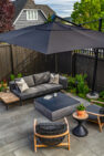 The fire pit area of a modern backyard with a generous square fire pit surrounded by an outdoor couch and two outdoor chairs. A large umbrella is open shading the fire pit and the fire pit is covered.