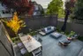 A overhead view of the patio of a refreshed backyard, paved in cast concrete pavers, and furnished with a dining table with room for six, a fire pit, and lounge furniture. Surrounding the perimeter of the patio are plantings.