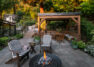 A single-family home’s updated yard showing a large patio of flagstone with lounge chairs upholstered in striped fabric near a lit fire pit featuring large gray spheres under the flames. Behind the fire pit is a wooden cabana with a grille, bench seating, and an outdoor dining area. Behind the cabana is a natural stone stair leading to the terraced yard beyond.