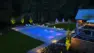 A backyard pool lit at night glows blue with pink accent lights. A hot tub, lounge areas, a fire pit, and kids play area are beyond. Woodinville Backyard Retreat – Residential Landscape Architecture – Board & Vellum