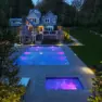 View of a new pool behind a large home, lit at night, glowing with blue water with pink accents in the pool and hot tub. Woodinville Backyard Retreat – Residential Landscape Architecture – Board & Vellum